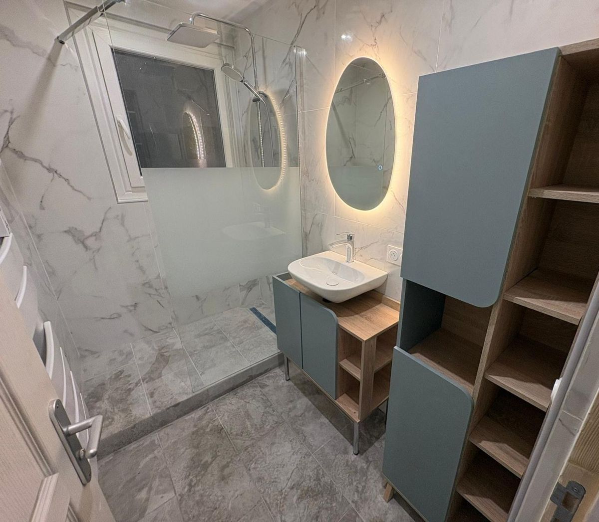 Salle de bains en carrelage effet marbre, douche vitrée, miroir rond lumineux, meuble vasque
